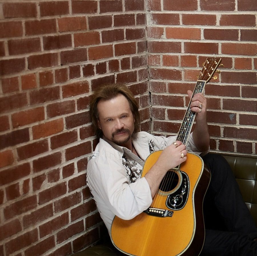 Travis Tritt sitting on a couch with his guitar.